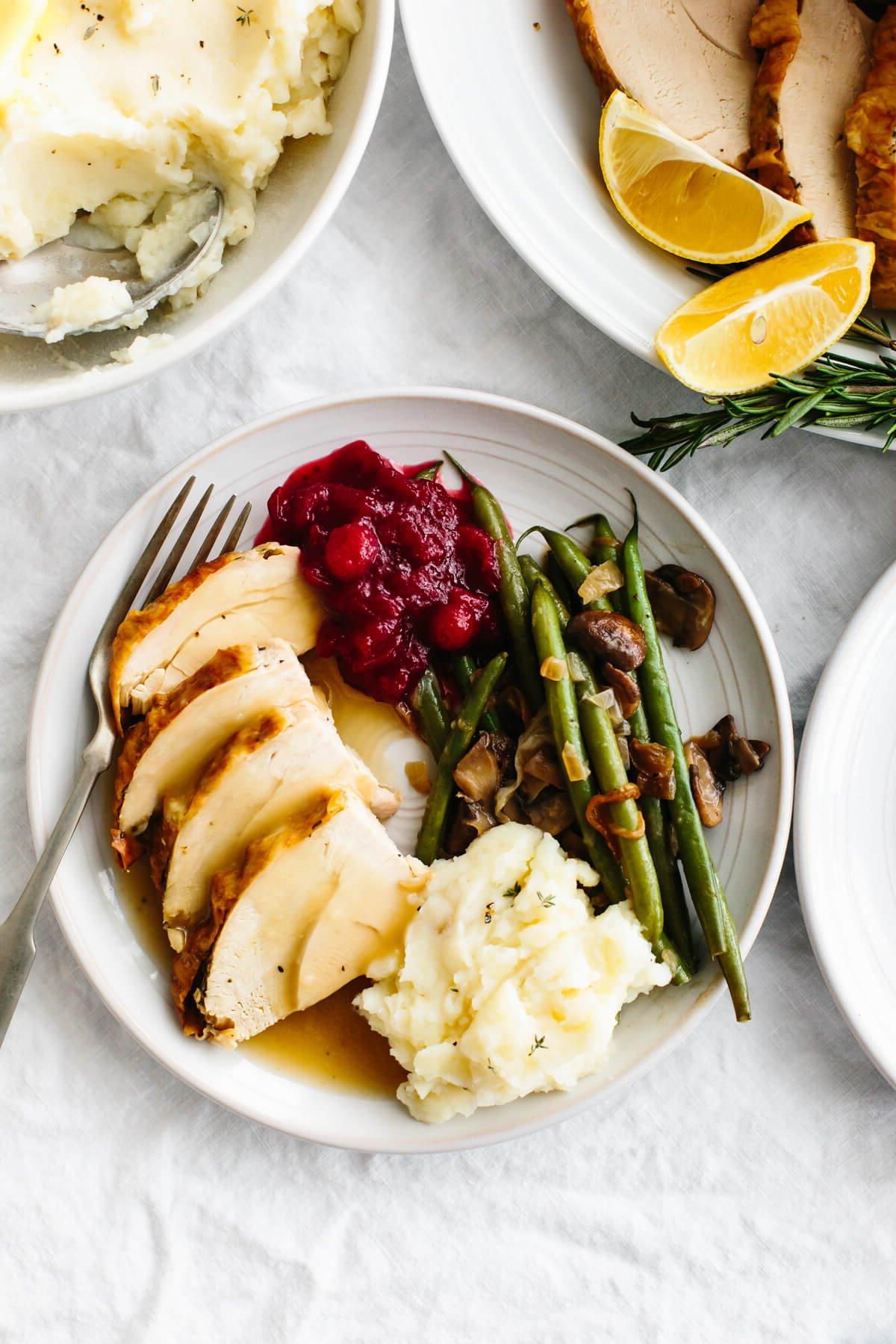 Dieses Thanksgiving-Menü bietet herbstliche Aromen, klassische Gerichte mit modernem Dreh und natürlich den ultimativen gebratenen Truthahn, den die ganze Familie lieben wird! Ein Teller mit Thanksgiving-Beilagen und gebratenem Truthahn.