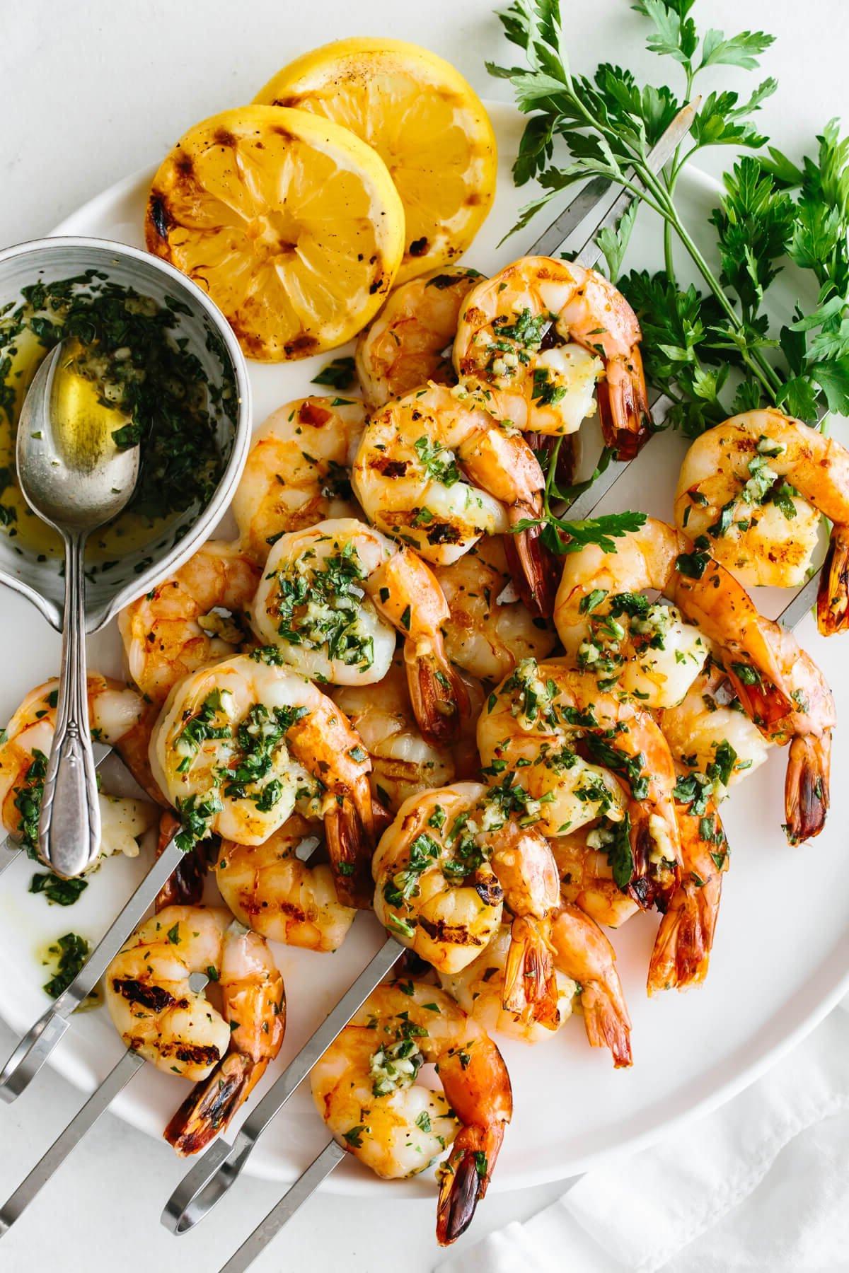 25+ Beste Memorial Day Rezepte 2 Teller mit gegrillten Garnelenspießen, garniert mit Zitronenscheiben, Petersilie und Marinade.