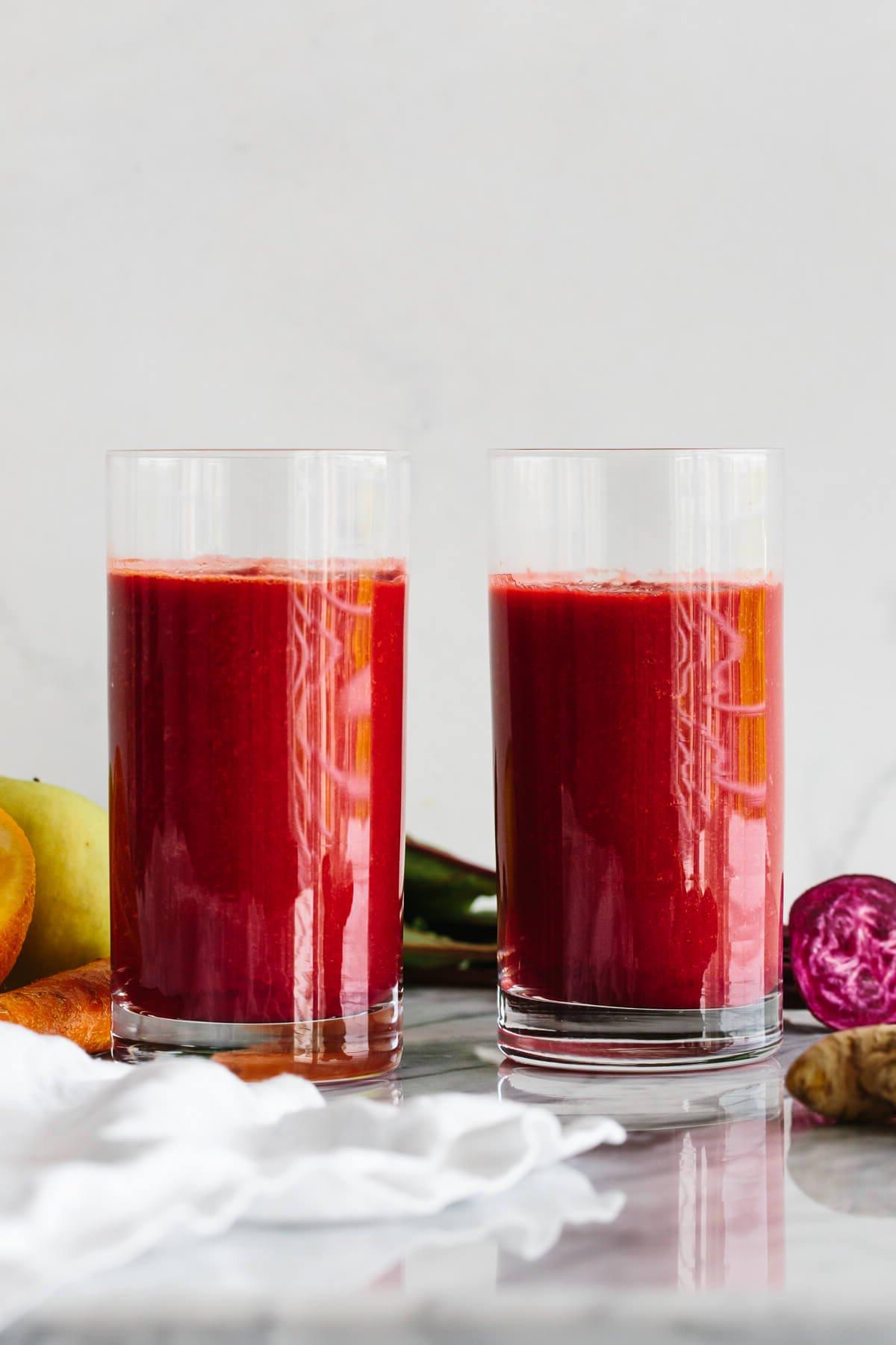 10+ Entzündungshemmende Getränke für mehr Wohlbefinden 11 Zwei Gläser Apfel-Karotten-Rote-Bete-Smoothie, umgeben von frischen Produkten.