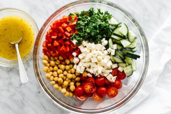 Zutaten für einen mediterranen Kichererbsensalat in einer Glasschüssel neben einer kleinen Schüssel Zitronenvinaigrette auf einem Tisch.