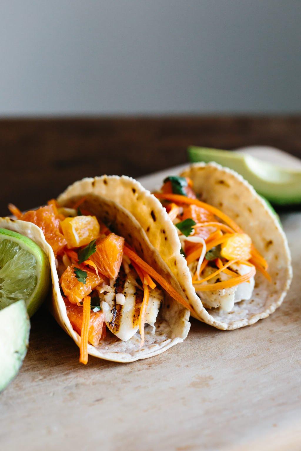 15+ einfache Grillrezepte für den Sommer 16 Zwei Fisch-Tacos auf einem Tisch neben Limetten und Avocado.