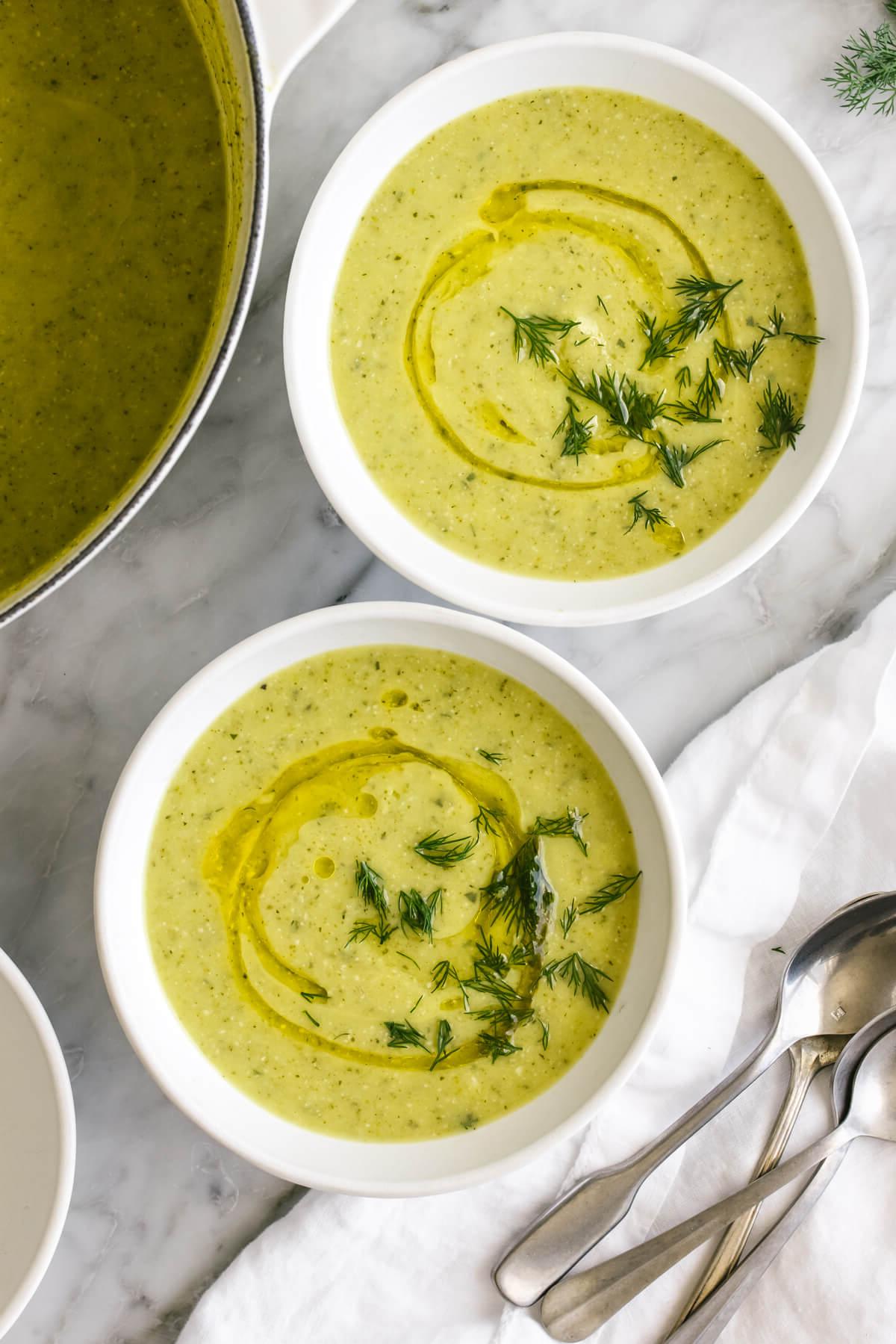Über 50 einfache Whole30 Abendessen-Rezepte 17 Zucchini-Suppe in zwei weißen Schüsseln auf einem Tisch neben Löffeln.