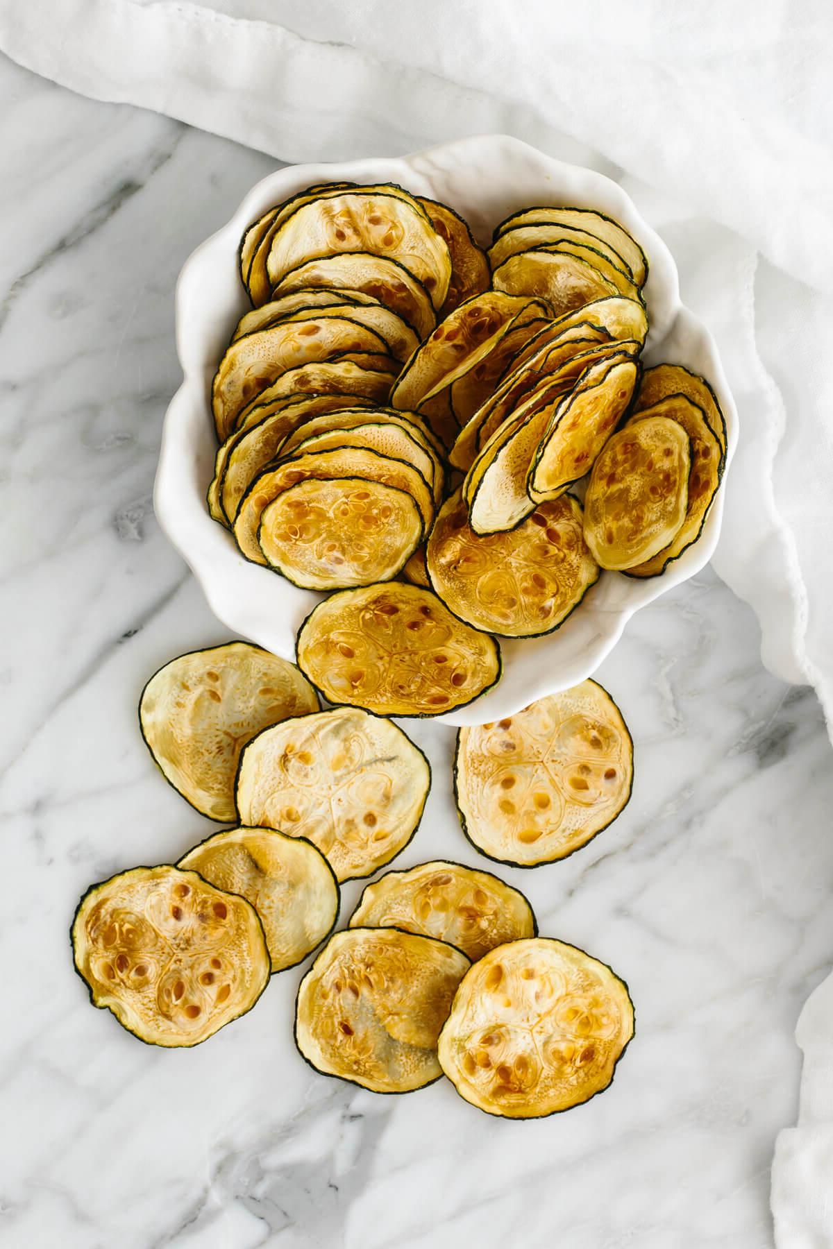 Über 20 beste Zucchini-Rezepte 18 Eine weiße Schüssel mit Zucchini-Chips und ein paar zusätzlichen Chips auf dem Tisch.