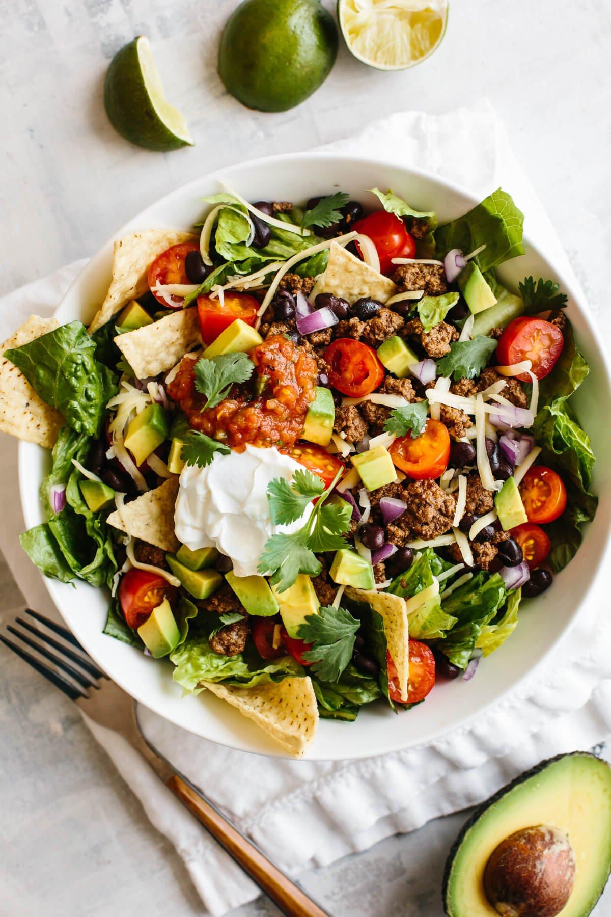 30 gesunde Rezepte in 30 Minuten oder weniger 26 Taco-Salat in einer weißen Schüssel auf einer Serviette, mit Limetten und einer halben Avocado neben der Schüssel.