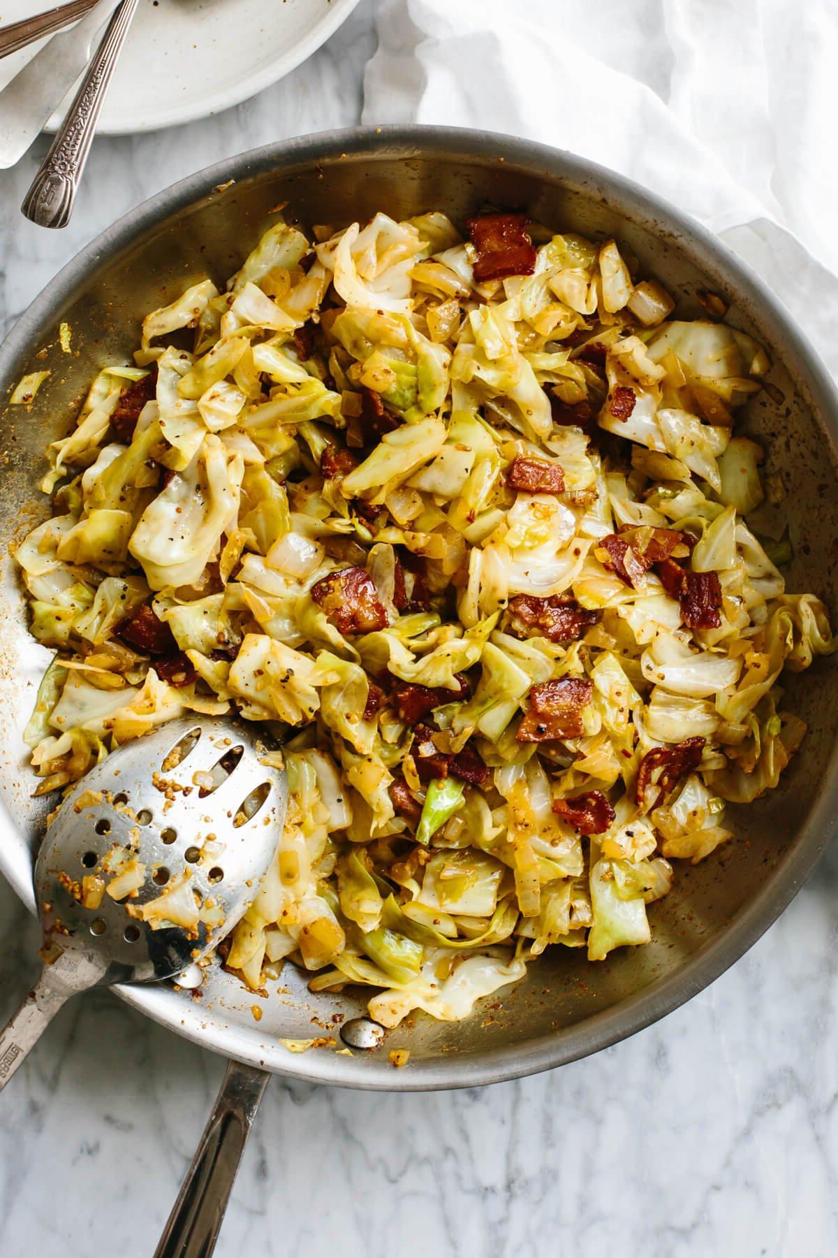 Mardi Gras Essensideen 6 Eine gusseiserne Pfanne mit gebratenem Kraut und knusprigem Speck, dazu ein großer Servierlöffel, appetitlich präsentiert auf einem Holztisch.
