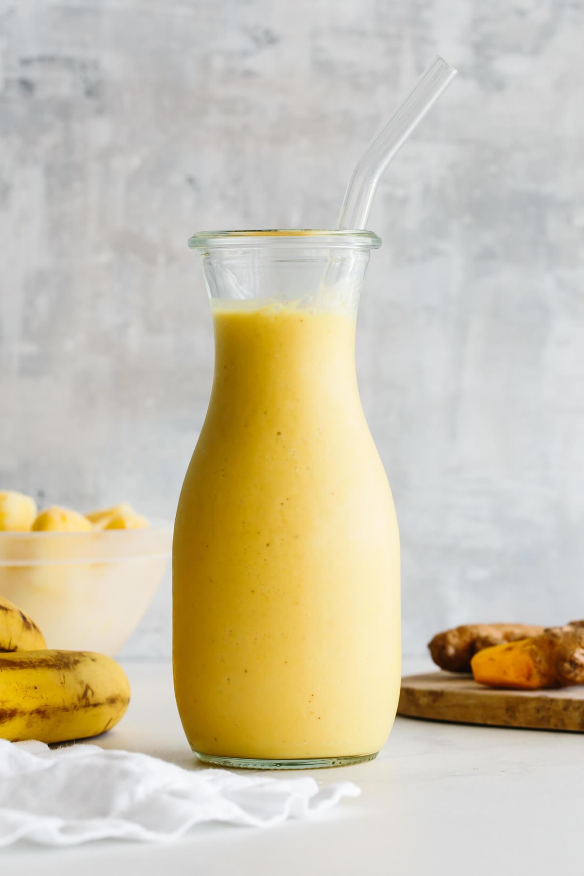 10+ Entzündungshemmende Getränke für mehr Wohlbefinden 7 Kurkuma-Smoothie in einem Glasgefäß mit Strohhalm und Zutaten im Hintergrund.