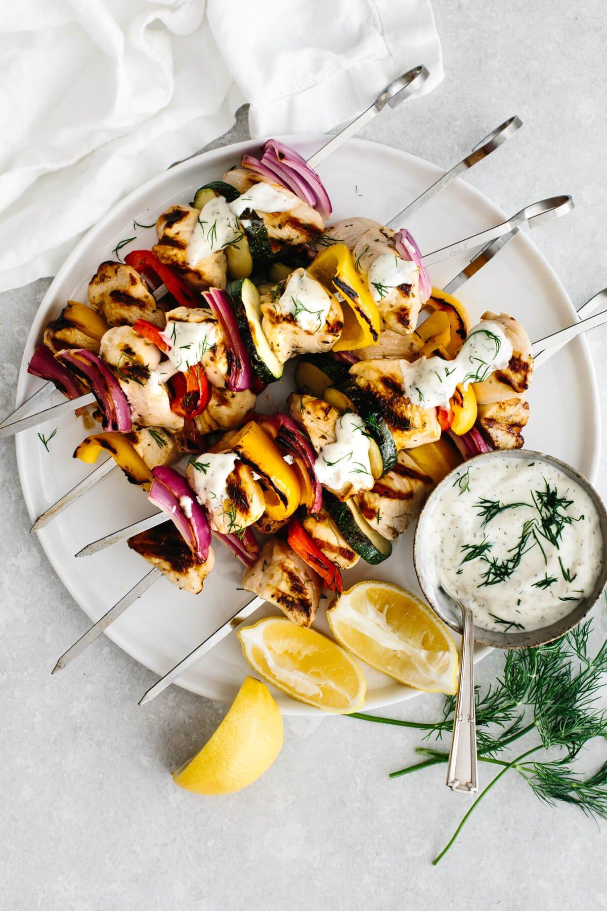 Griechische Hähnchenspieße auf einem weißen Teller mit Tzatziki-Sauce.