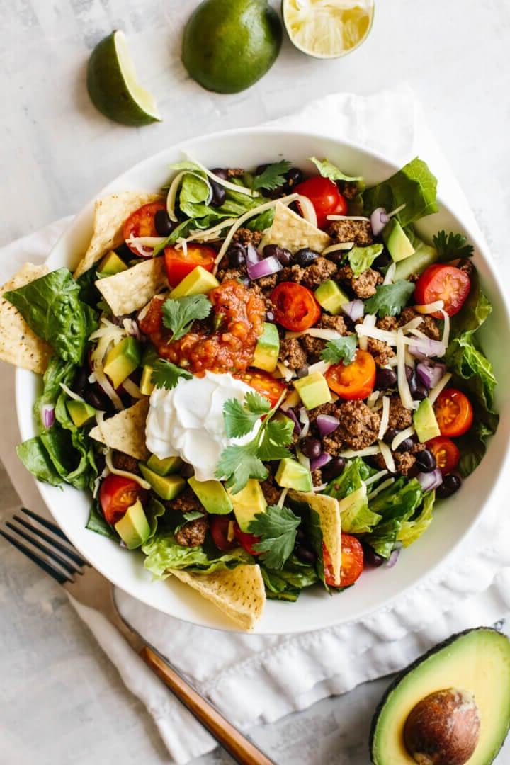 Taco-Salat in einer weißen Schüssel auf einer Serviette, mit Limetten und einer halben Avocado neben der Schüssel.