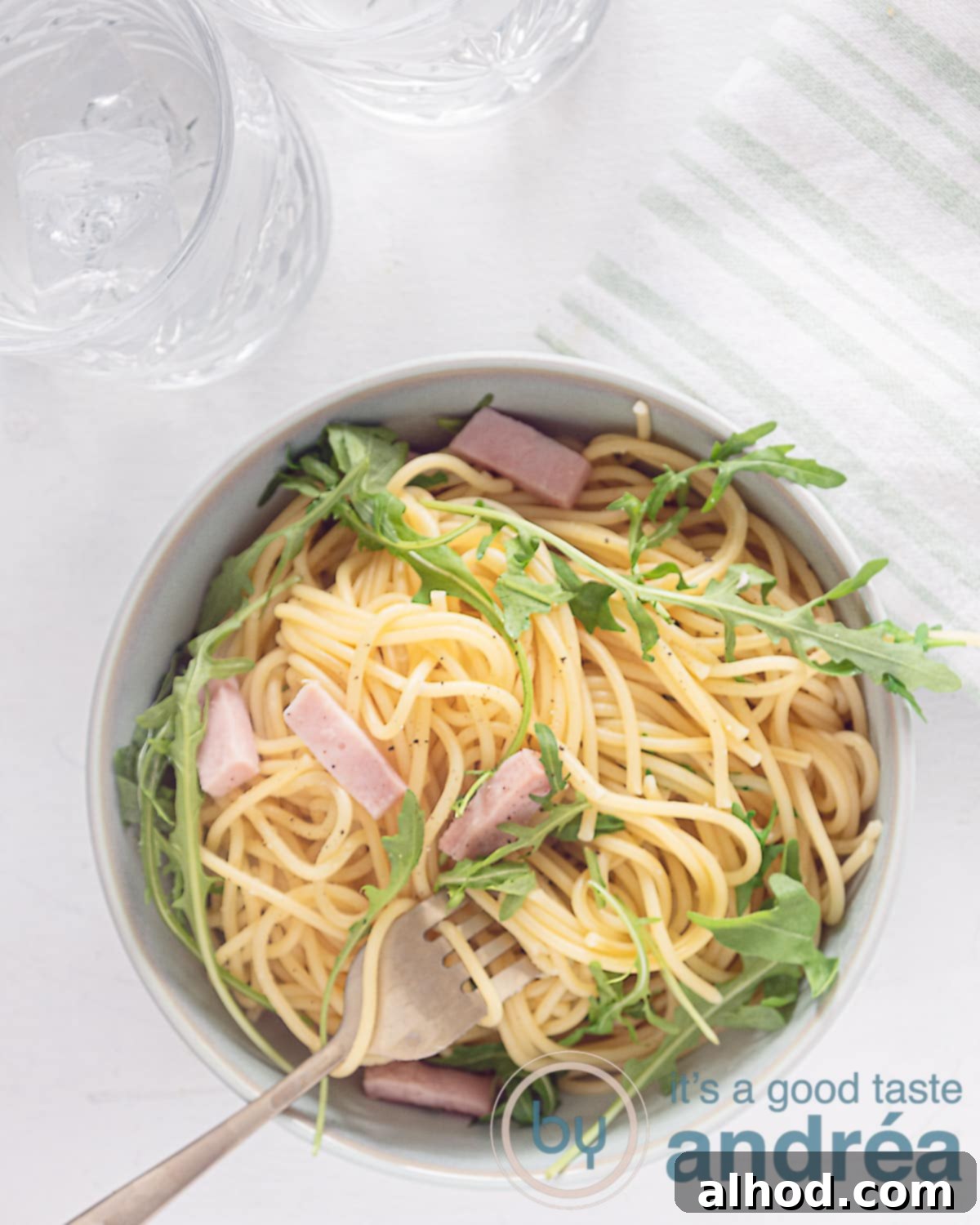 Hearty Italian Ham Spaghetti 2 A photo from above of a white background with a bowl filled with spaghetti, ham and arugula. A golden fork with spaghetti curled up, ready to be eaten.