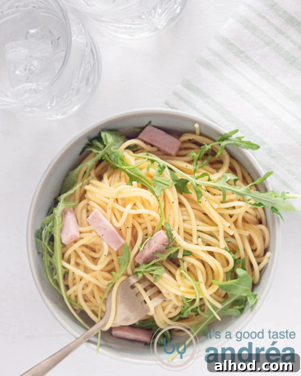Classic Italian Ham Spaghetti 2 A photo from above of a white background with a bowl filled with spaghetti, ham and arugula. A golden fork with spaghetti curled up, ready to be eaten.