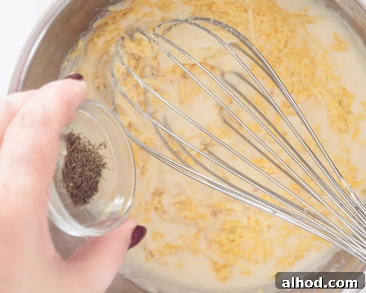 Effortless Cheesy Cod Bake 5 A creamy white sauce in a saucepan, being stirred with a whisk, with fresh green dill sprigs added for flavor.