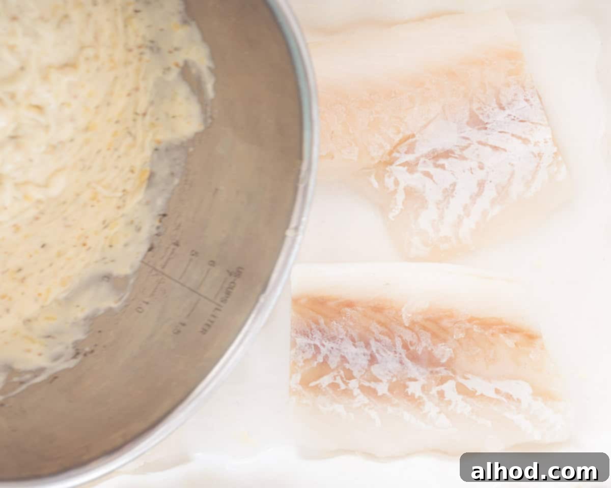Effortless Cheesy Cod Bake 6 A white casserole dish prepared with four cod fillets, over which a creamy white sauce is being poured evenly, covering the fish.
