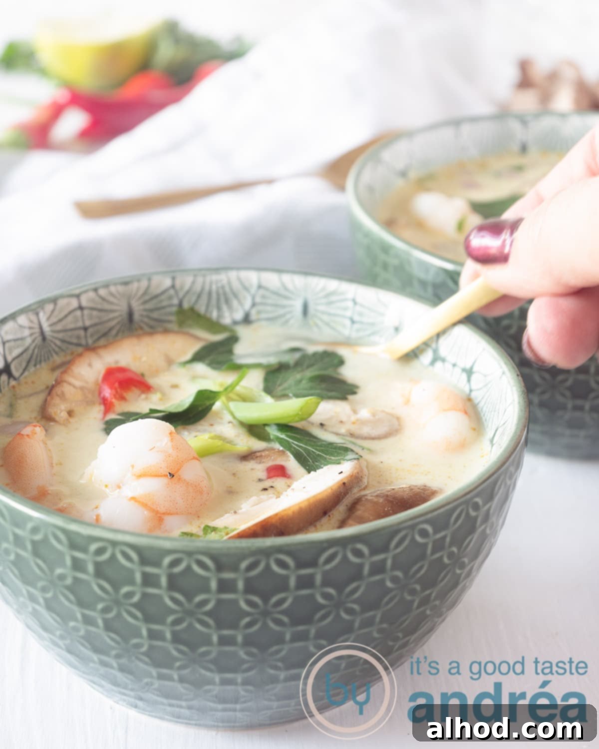 Tom Kha Goong Thai Coconut Shrimp Soup 5 A green bowl filled with Thai coconut soup, shiitake and shrimp. A hand taking a spoon. A second bowl in the back