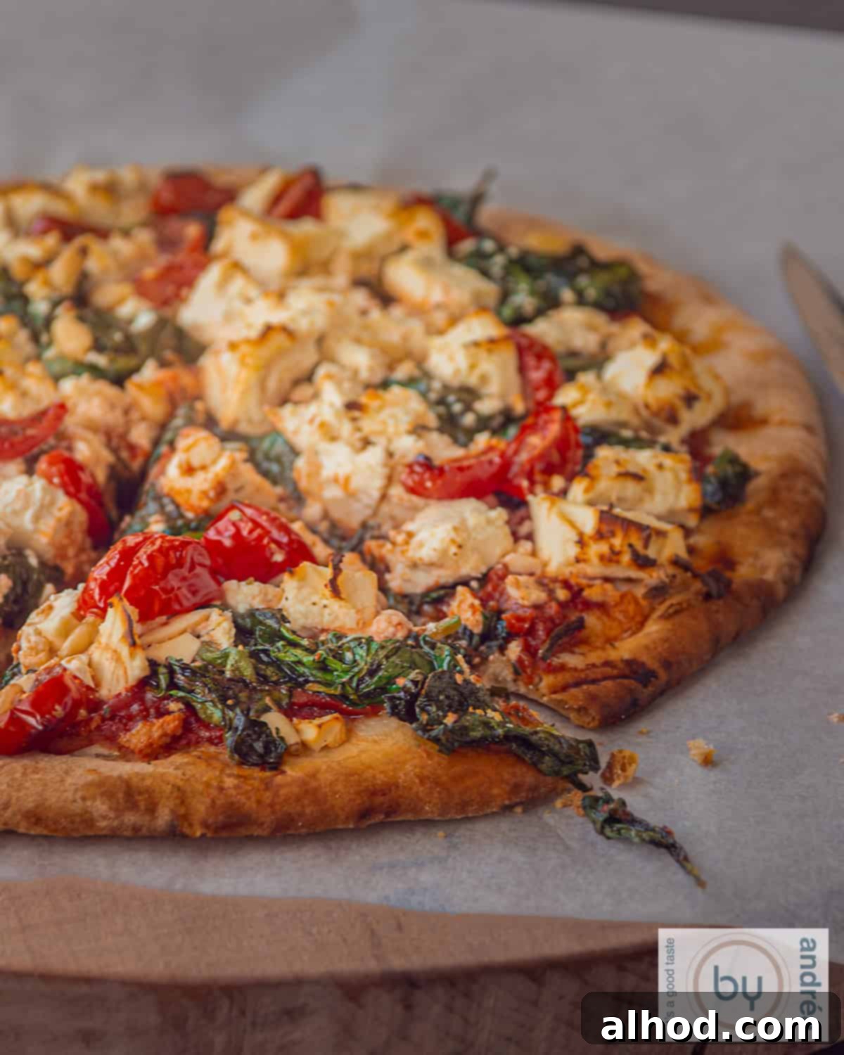 Artisan Spinach Feta Pizza with Garden Tomato Sauce 2 A slice of pizza with feta, spinach, tomato and pine nuts on a wooden board on baking paper.