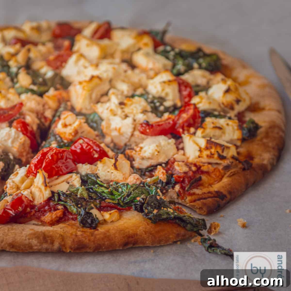 Artisan Spinach Feta Pizza with Garden Tomato Sauce 4 A square photo of a pizza, from which a slice has been cut, on white baking paper. The pizza is topped with feta, tomato, spinach, and pine nuts.