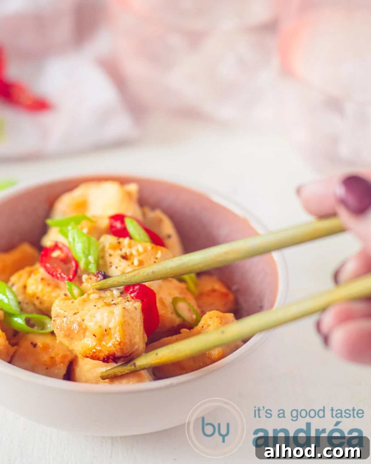 Ginger-Glazed Honey Chicken 5 A height photo with a pink container filled with chicken cubes with a honey-ginger glaze, spring onion and red pepper. Two gold chopsticks grab a chicken cube. A white background. In the background a pink white tea towel and a pink water glass.