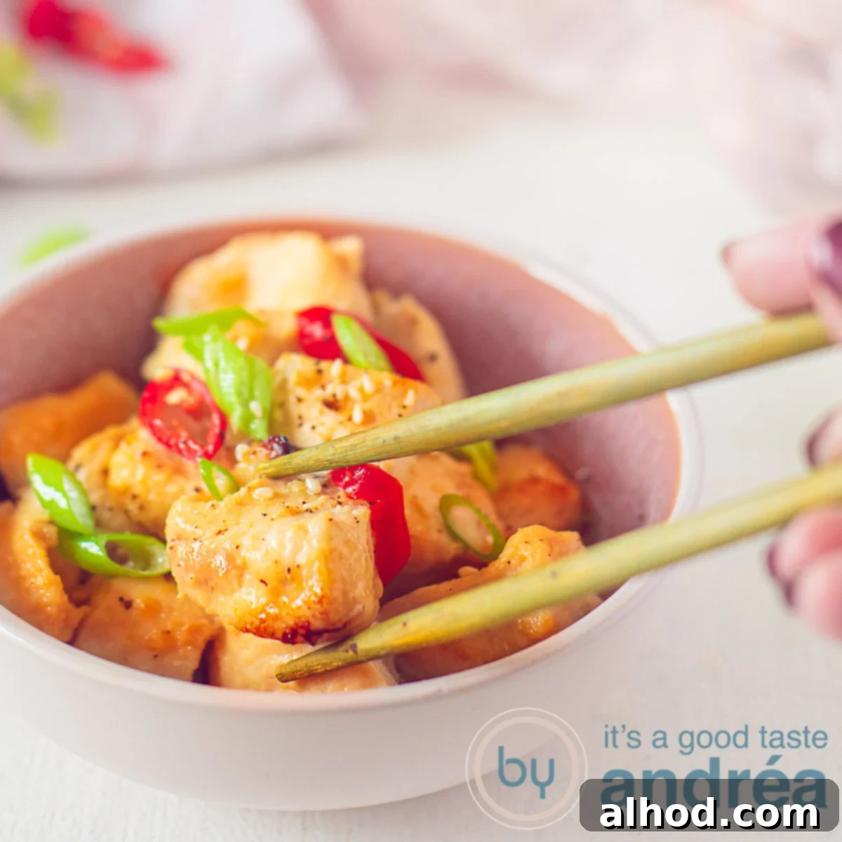 Ginger-Glazed Honey Chicken 6 A square photo with a pink dish with honey ginger chicken, spring onion and pepper. Two chopsticks grab a chicken cube. In the background a pink and white tea towel.