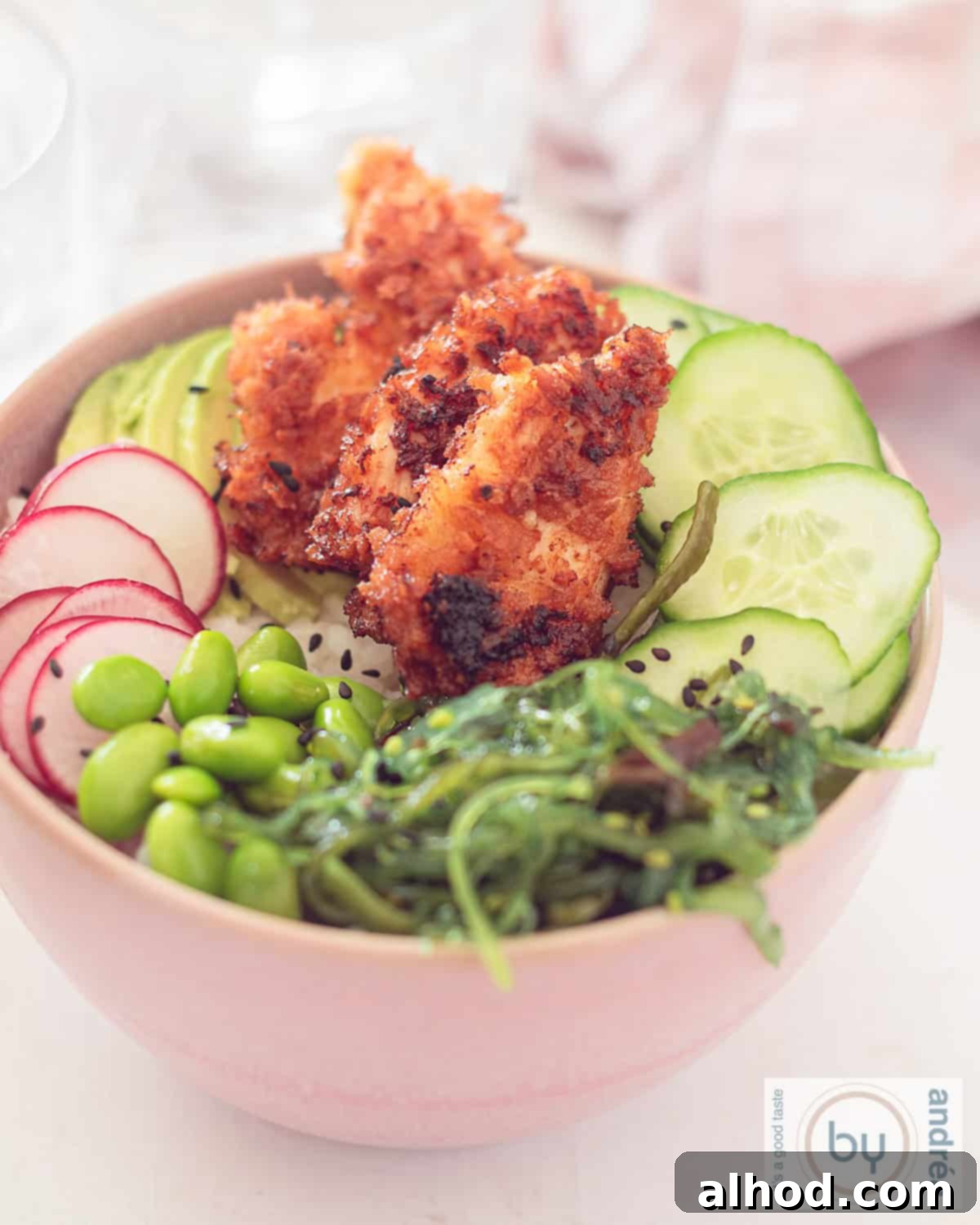 25-Minute Crispy Sriracha Chicken Sushi Bowl 2 A shot of a pink poke bowl filled with edamame beans, radish, seaweed salad, cucumber, and crispy sriracha chicken on a white background.