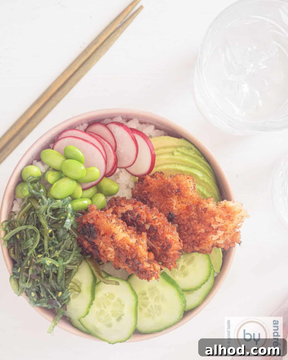 25 Minute Crunchy Sriracha Chicken Sushi Bowl 3 Overhead shot of a pink poke bowl with crispy chicken, seaweed salad, edamame, cucumber, radish, and avocado on a white background.