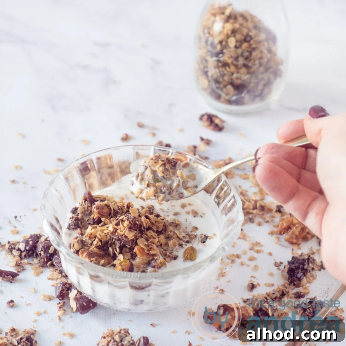 7 Scrumptious Mother's Day Brunch Recipes She'll Love 4 A square photo featuring a glass bowl filled with creamy yogurt and crunchy dark chocolate granola, with a golden spoon taking a bite, set on a white marble background. A glass jar of more chocolate nut granola is visible in the background.