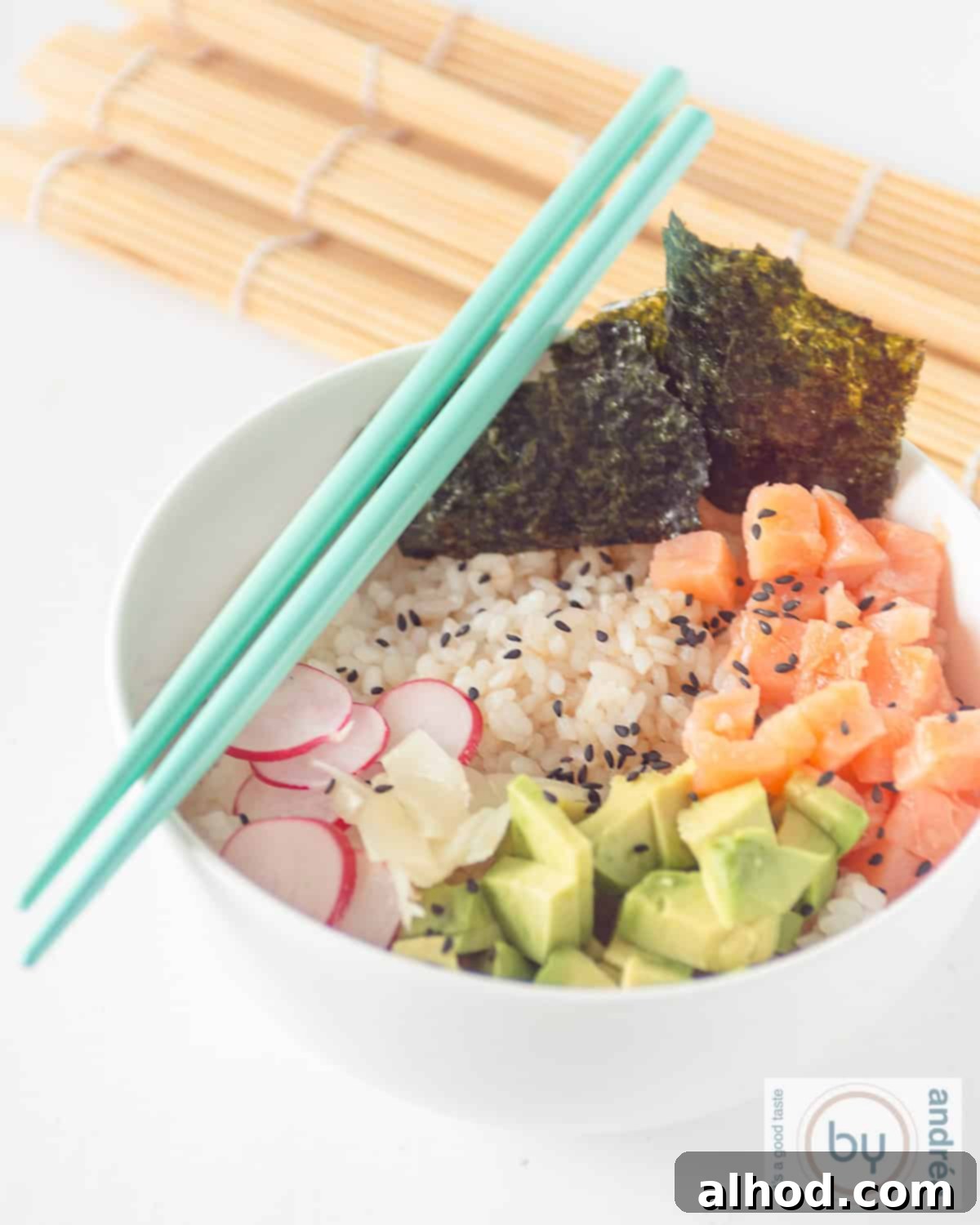Simple Smoked Salmon Sushi Bowl Recipe 2 A white background with a white bowl filled with smoked salmon, sushi rice, avocado, radish, and nori. Two blue chopsticks lie on the bowl.