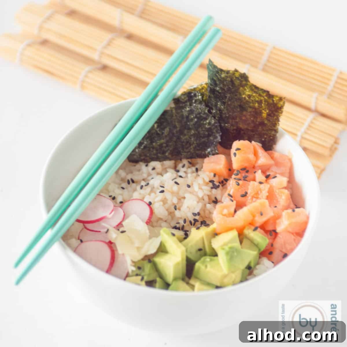Homemade Smoked Salmon Sushi Bowl 4 A square photo of a white bowl with sushi rice, radish, avocado, smoked salmon, and seaweed. Two blue chopsticks and a wicker mat decorate the table.