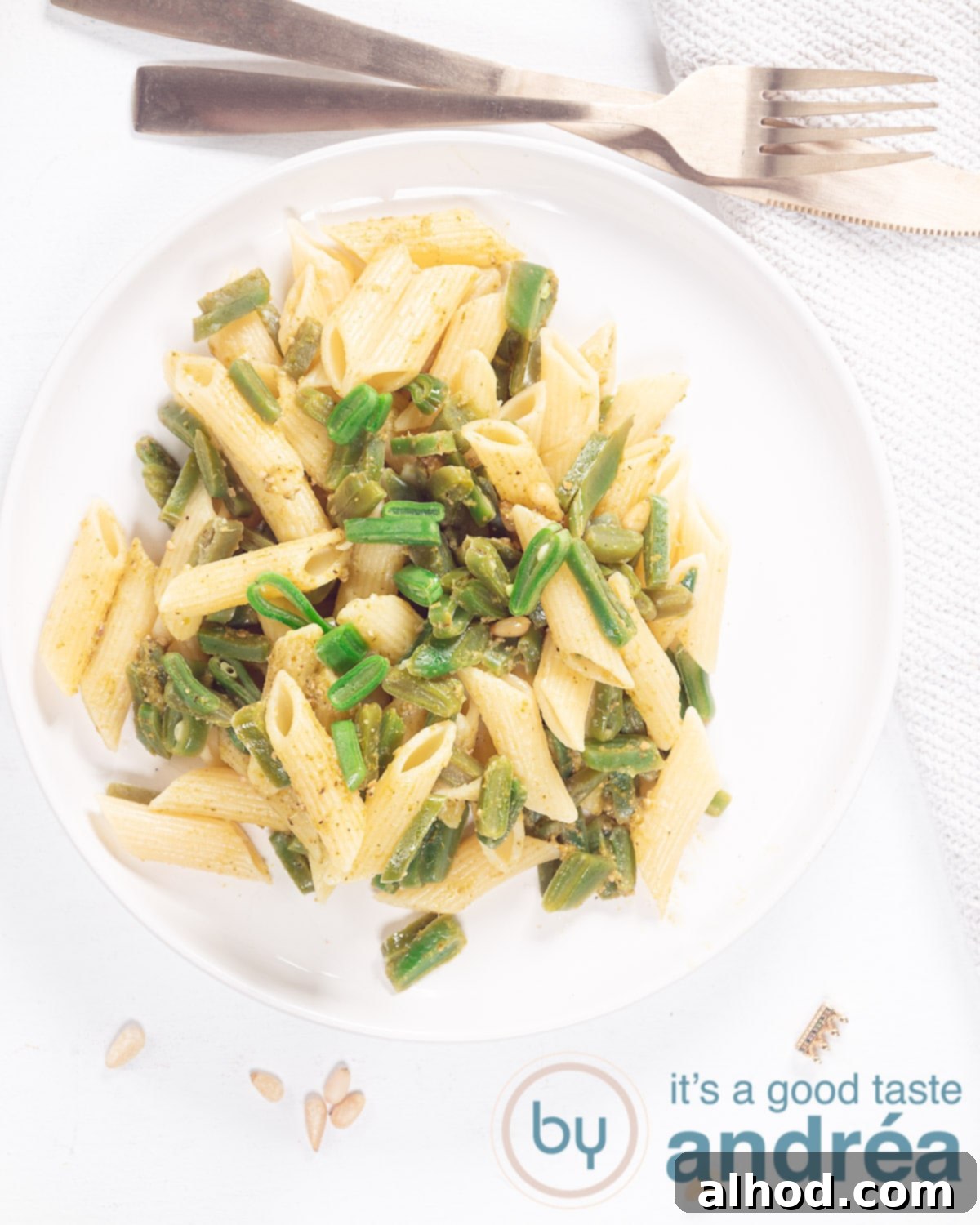 Fresh Runner Bean Pesto Pasta 2 A photo from above: a white plate filled with penne, string beans and pesto on a white background. Above it a golden fork and knife.