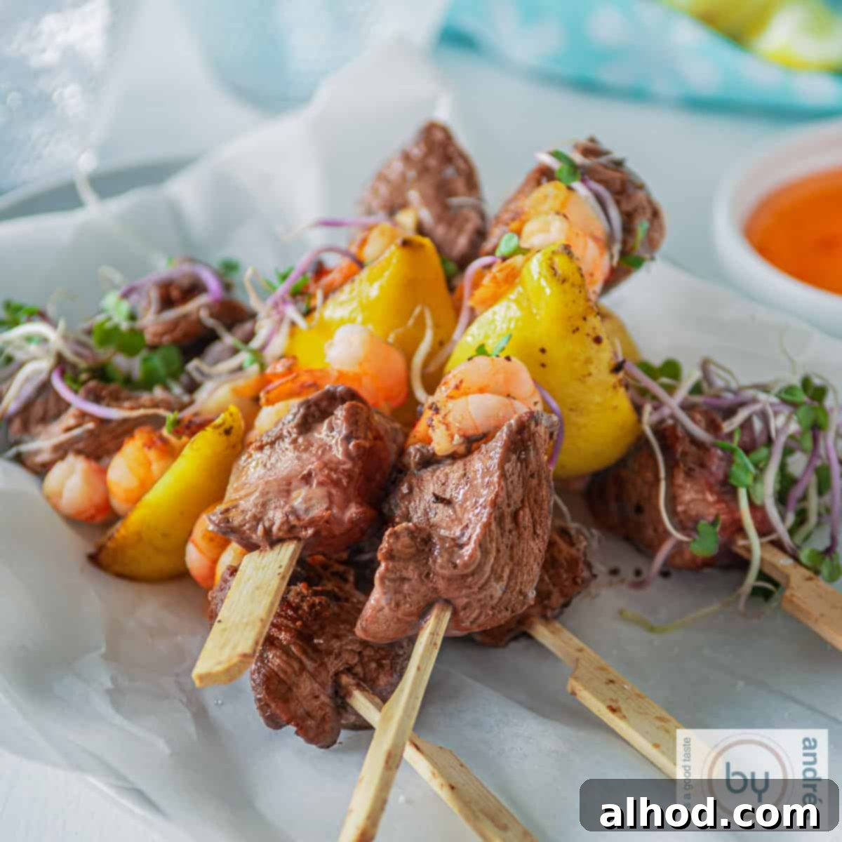 Grilled Surf and Turf Skewers 4 A square photo with a white background featuring five skewers of surf and turf steak with shrimp. A bowl of chili sauce and a blue cloth are visible in the background, suggesting a complete meal.