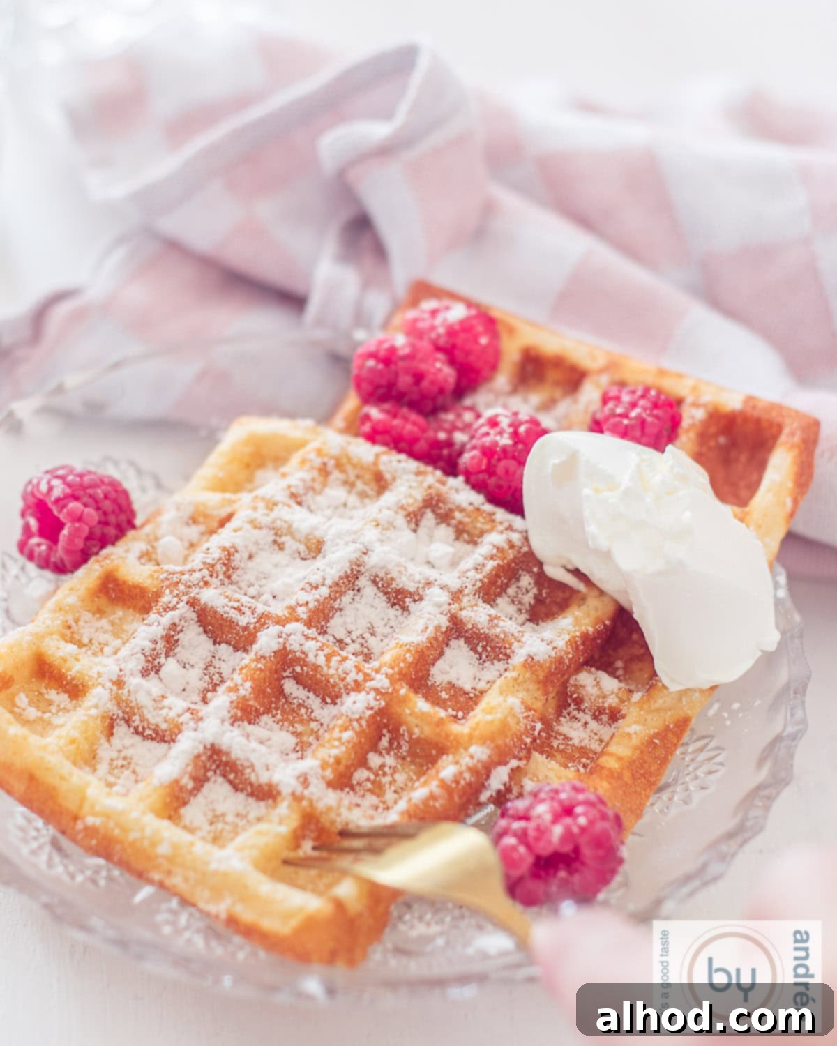 A close-up photograph showcasing two beautifully golden-brown waffles on a pristine white glass plate, lightly dusted with confectioner's sugar and generously topped with vibrant fresh raspberries and a rich dollop of mascarpone, presented against a soft, unfocused white background.