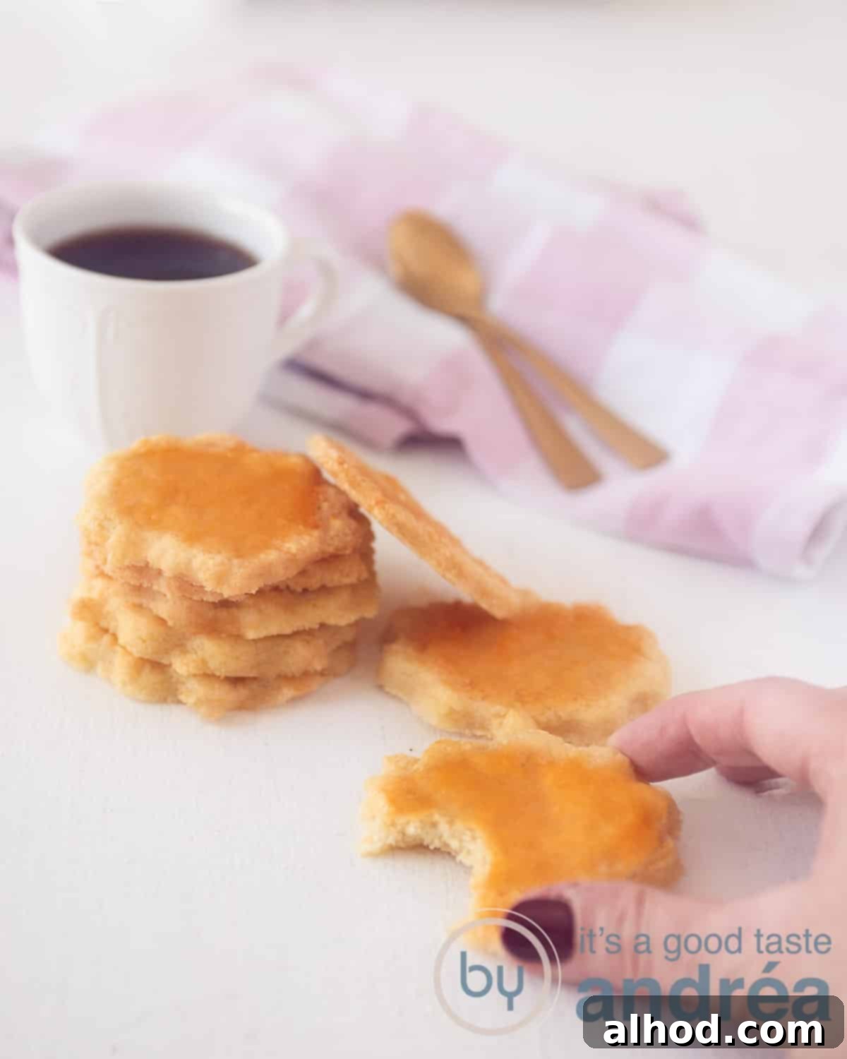 Delicate French Butter Sables 5 A close-up shot of five delectable sable cookies stacked neatly on top of each other, with two more leaning invitingly to the side. In the foreground, a cookie with a perfect bite taken out of it is held by a hand, suggesting its irresistible nature. The background features a soft white and pink towel and a steaming cup of coffee, creating a cozy and inviting atmosphere for enjoying these melt-in-your-mouth French treats.