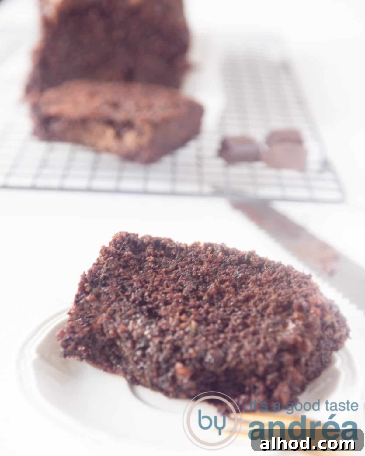 Decadent Dark Chocolate Zucchini Cake 5 A height photo with a white plate with a slice of zuchhini pound cake with chocolate in front. In the back the remaining loaf on a rack