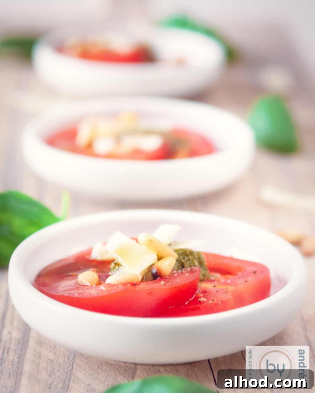 A height photo of a wooden board with three white bowls with tomato, cheese, pine nuts and pesto. Basil leaves are around it