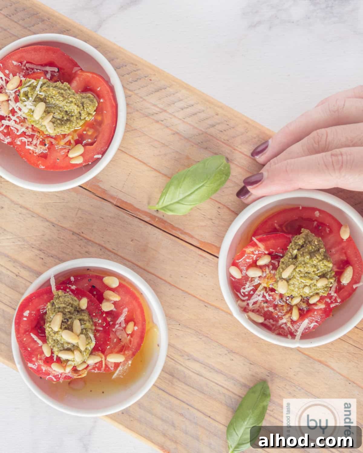 A height photo from above of a wooden board with three bowls of tomato slices, pesto, cheese and pine nuts. A hand takes a bowl.