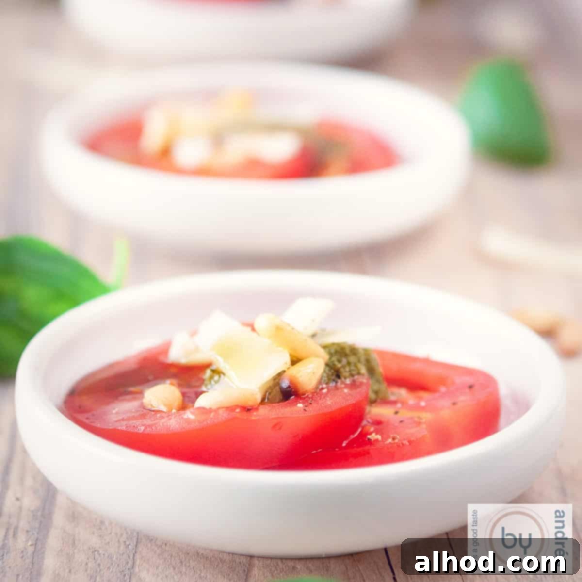 A square photo with a wooden background with three white bowls filled with tomato slices, grated cheese, pesto and pine nuts. Cheese and basil are around it.