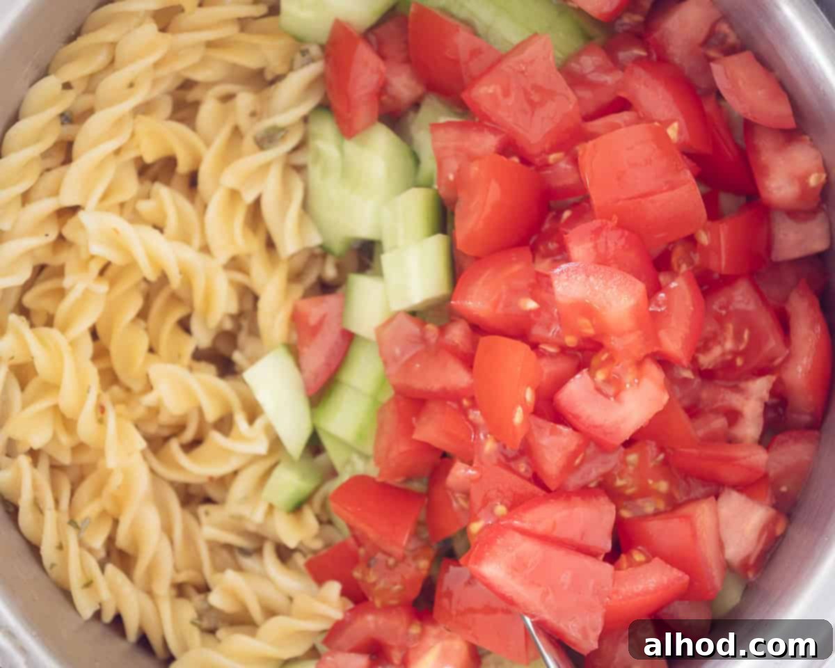 A bowl with cooked fussili, dressing, cucumber and tomato cubes