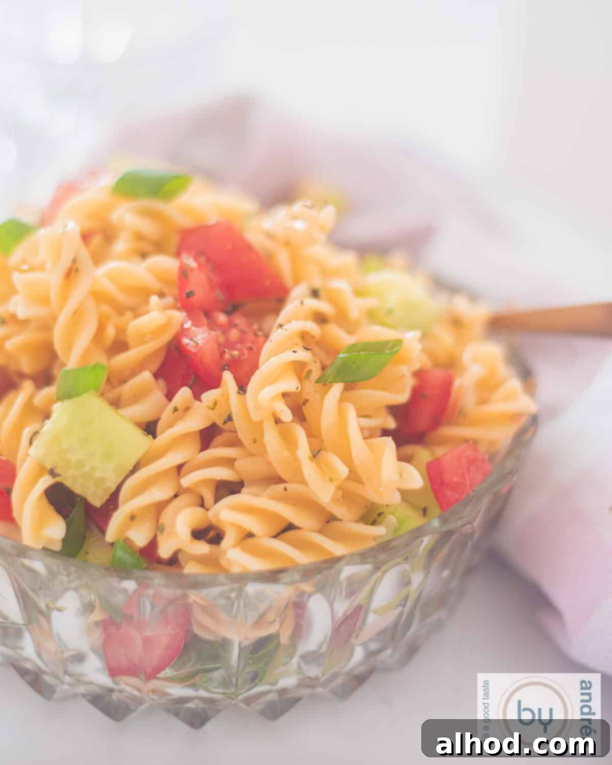 Zesty Italian Veggie Pasta Salad 9 A glass bowl filled with fusilli, cucumber, spring onions, tomatoes and dressing. A pink/white towel in the back.