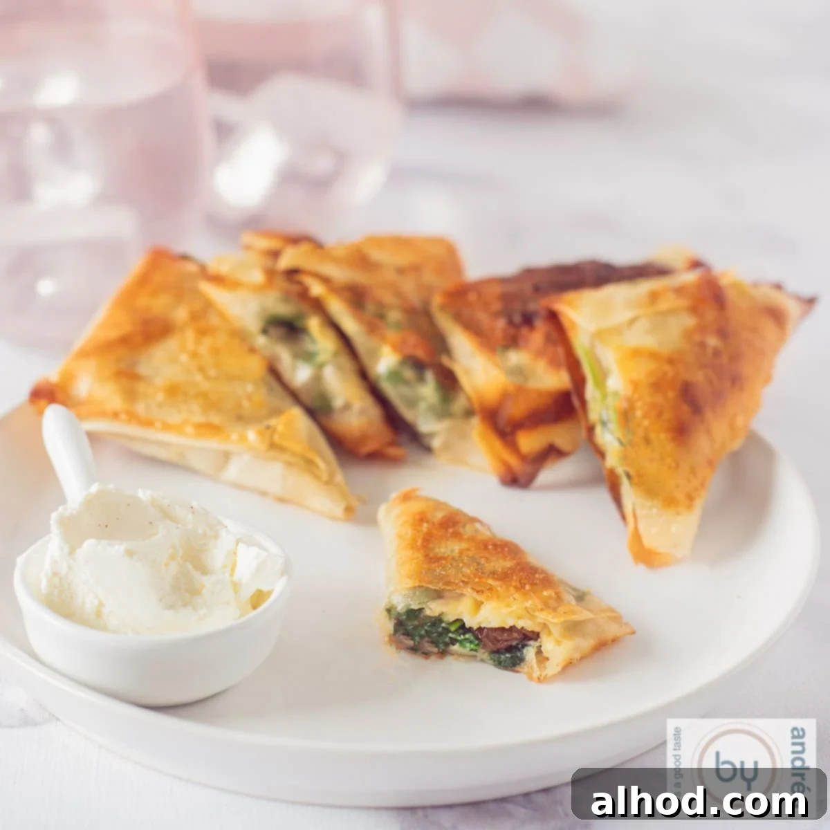 Crispy Spinach and Feta Bites 4 A square photo of a white plate with filo pastry spinach snacks in a row. A dip is diagonally in front of it. One of the snacks is halved so you can see the filling.