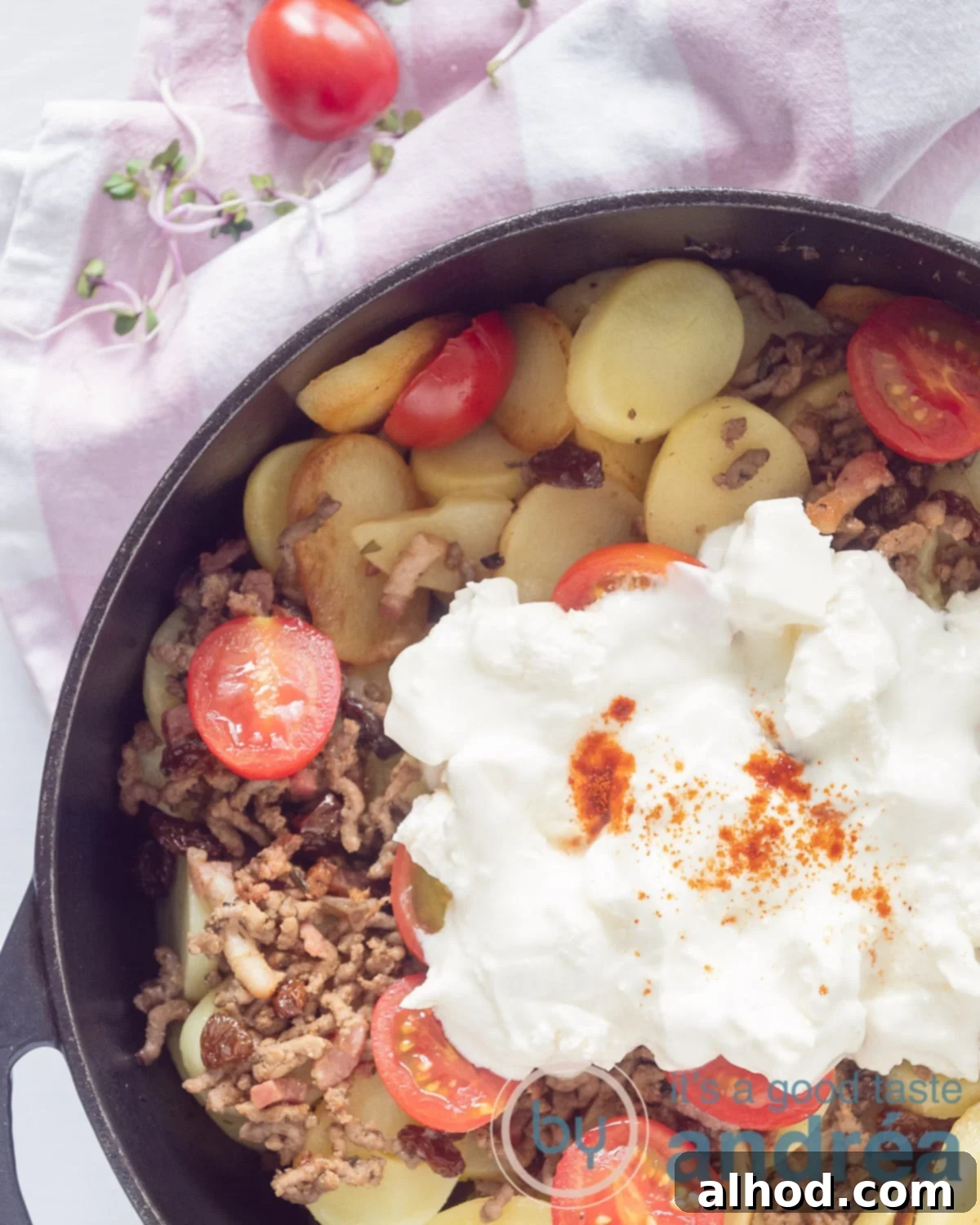 Hearty Greek Beef and Potato Bake 2 A photo from above of a black skillet filled with potatoes, tomatoes, zucchini, minced meat and a yogurt cheese topping. Above the pan a pink white tea towel.
