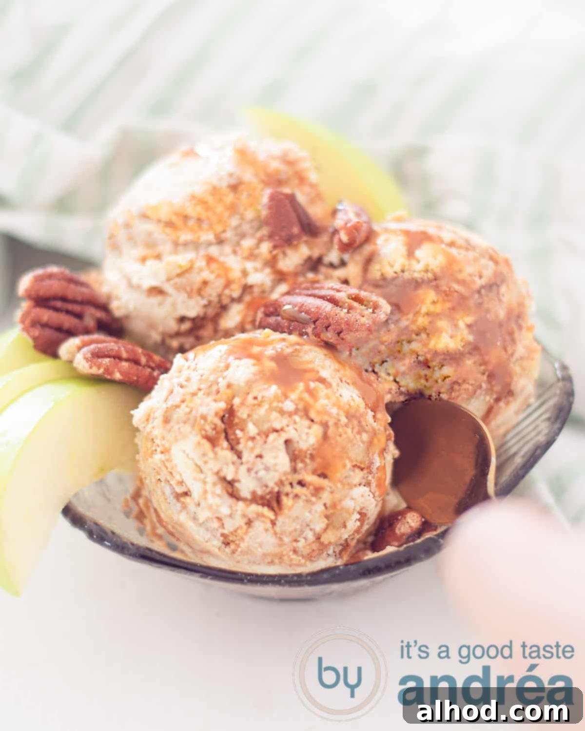 Easy Apple Pie Ice Cream 2 An elevation photo with a bowl filled with three scoops of ice cream with apples, apple wedges, pecans on a white background. A green and white tea towel in the background