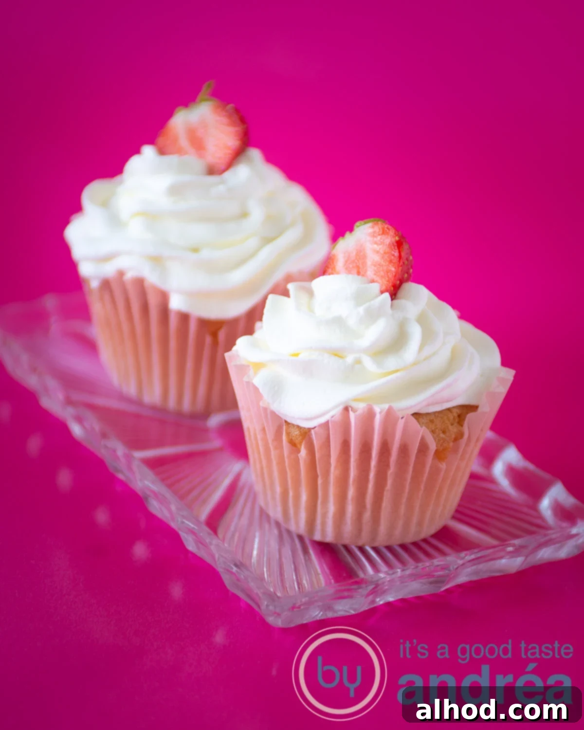 Strawberry Burst Cupcakes 2 Two perfectly decorated strawberry filled cupcakes with whipped cream and fresh strawberry halves, arranged on a glass plate.