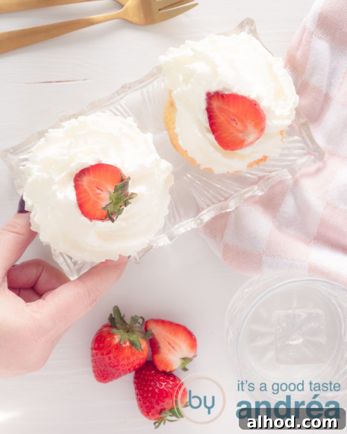 Sweet Strawberry Bliss Cupcakes 5 A photo from above with two strawberry cupcakes garnished with fresh strawberry slices. A hand is reaching for a cupcake from a glass bowl, with more strawberries scattered below.