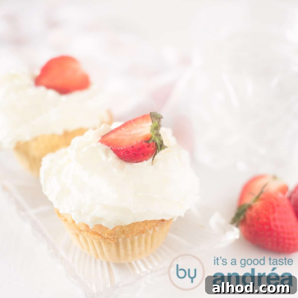 Strawberry Burst Cupcakes 6 A square photo of two cupcakes on a white surface on a bowl with a dollop of whipped cream and a strawberry on top