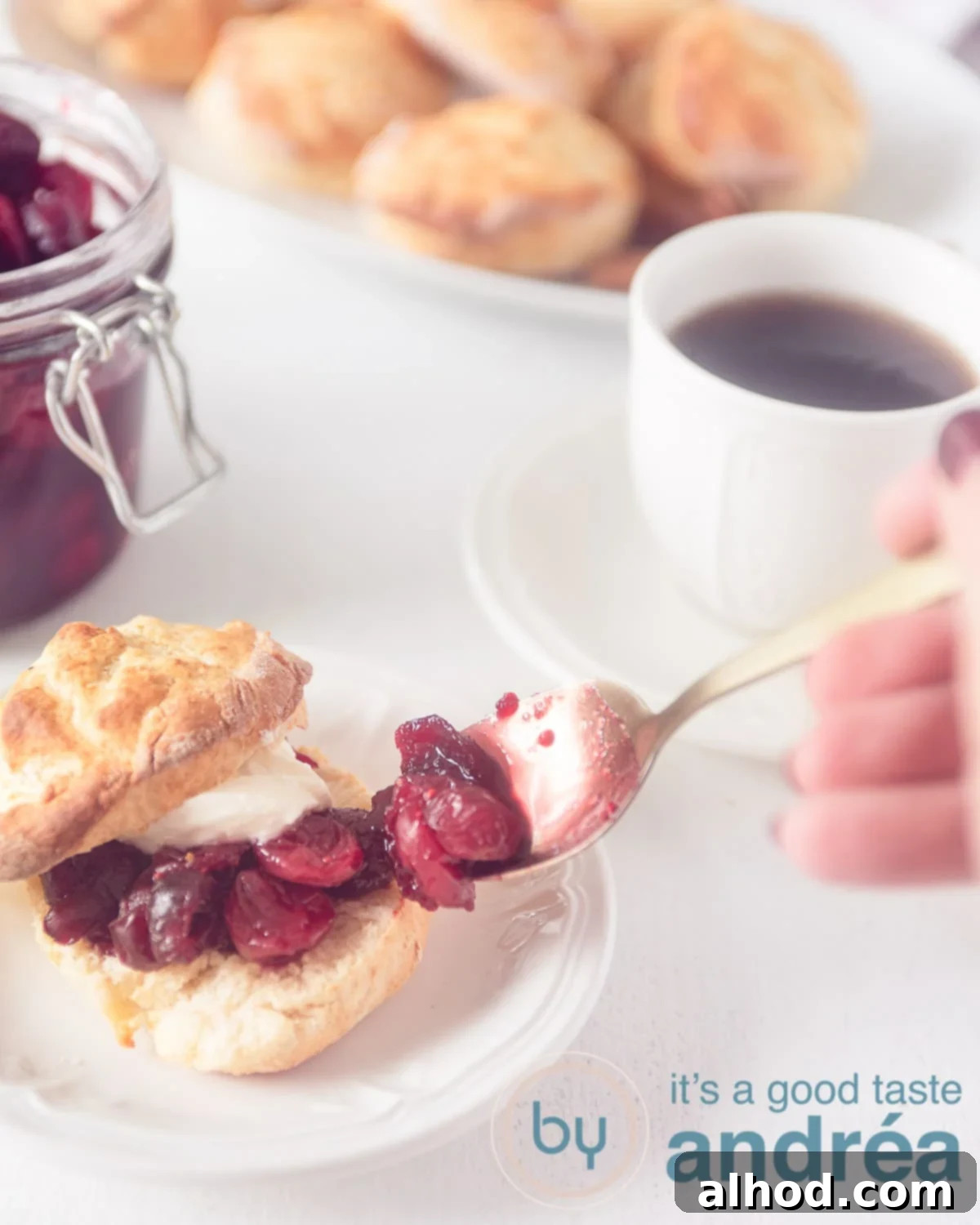 Fluffy Scones with Bright Cranberry Compote 2 A hand putting extra cranberry compote on a cut open scone. In the background scones, a glass jar with cranberry jam and a cup of coffee