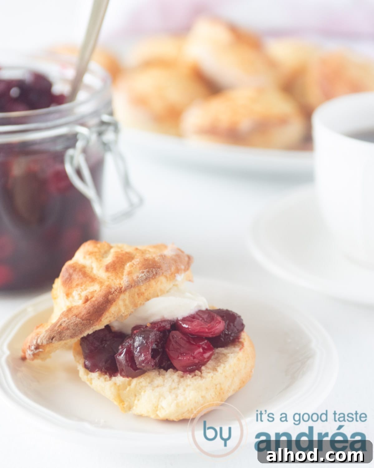 Fluffy Scones with Bright Cranberry Compote 5 a sliced scone on a white plate topped with cranberry compote and clotted cream on a white background. In the back a jar of cranberry compote, scones and a cup of tea