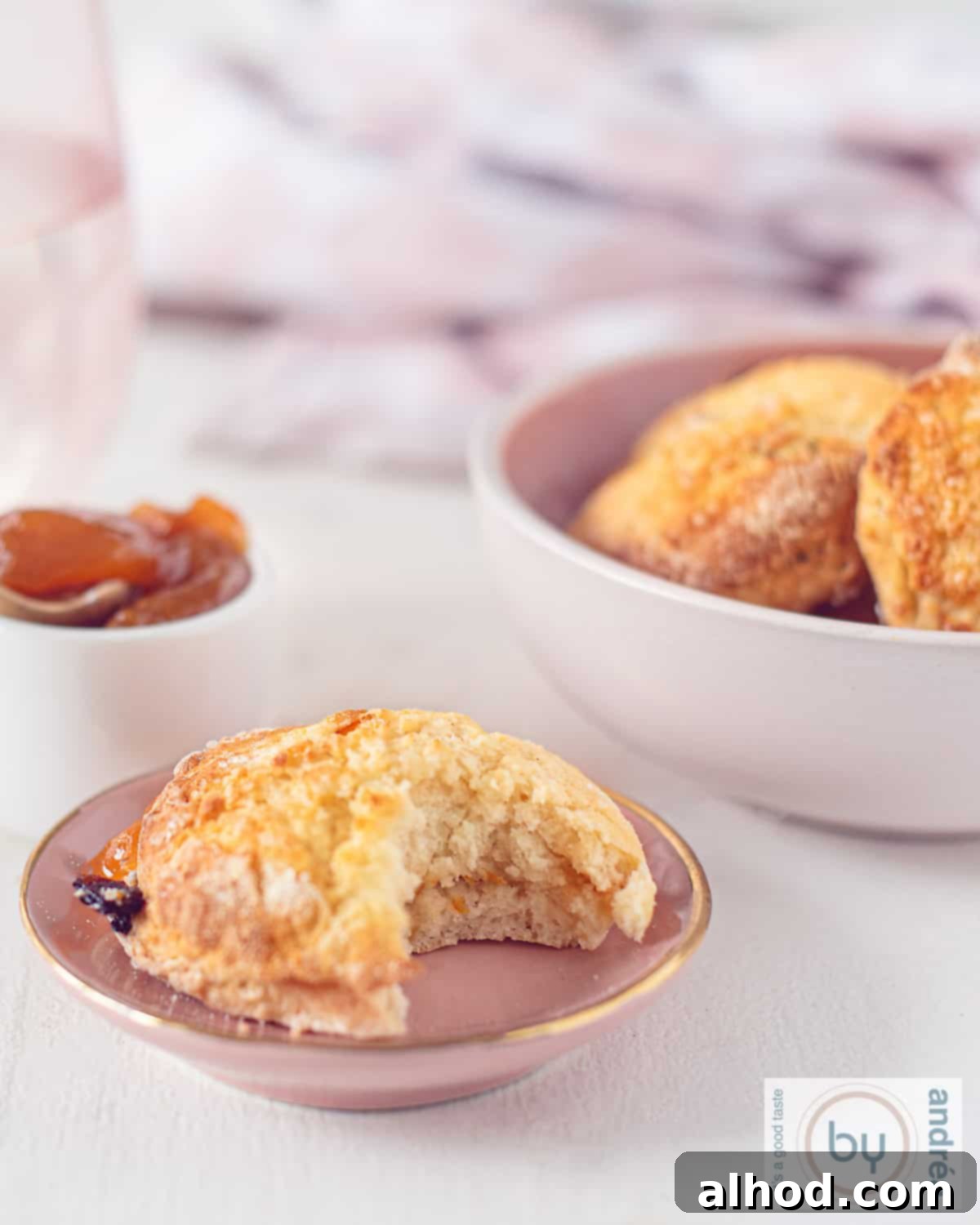 A height picture with currant orange scones, a bite taken out, showing the fluffy inside. A bowl with more scones in the back and some orange jam.