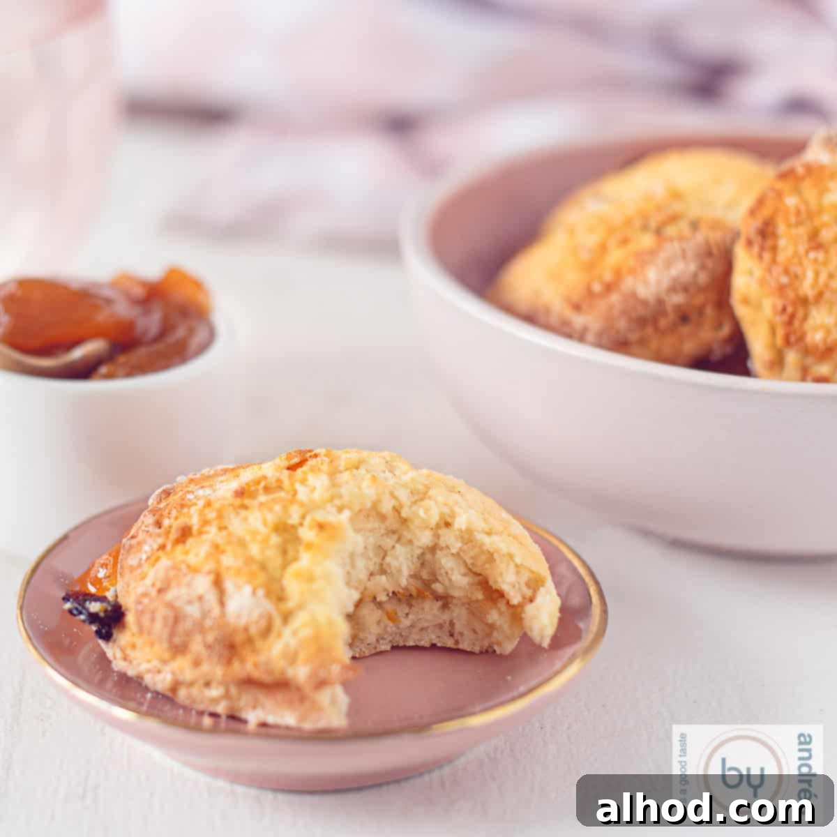 A square picture of a pink plate with a scone, a bite taken out of it. In the back a bowl with more scones and some orange jelly.