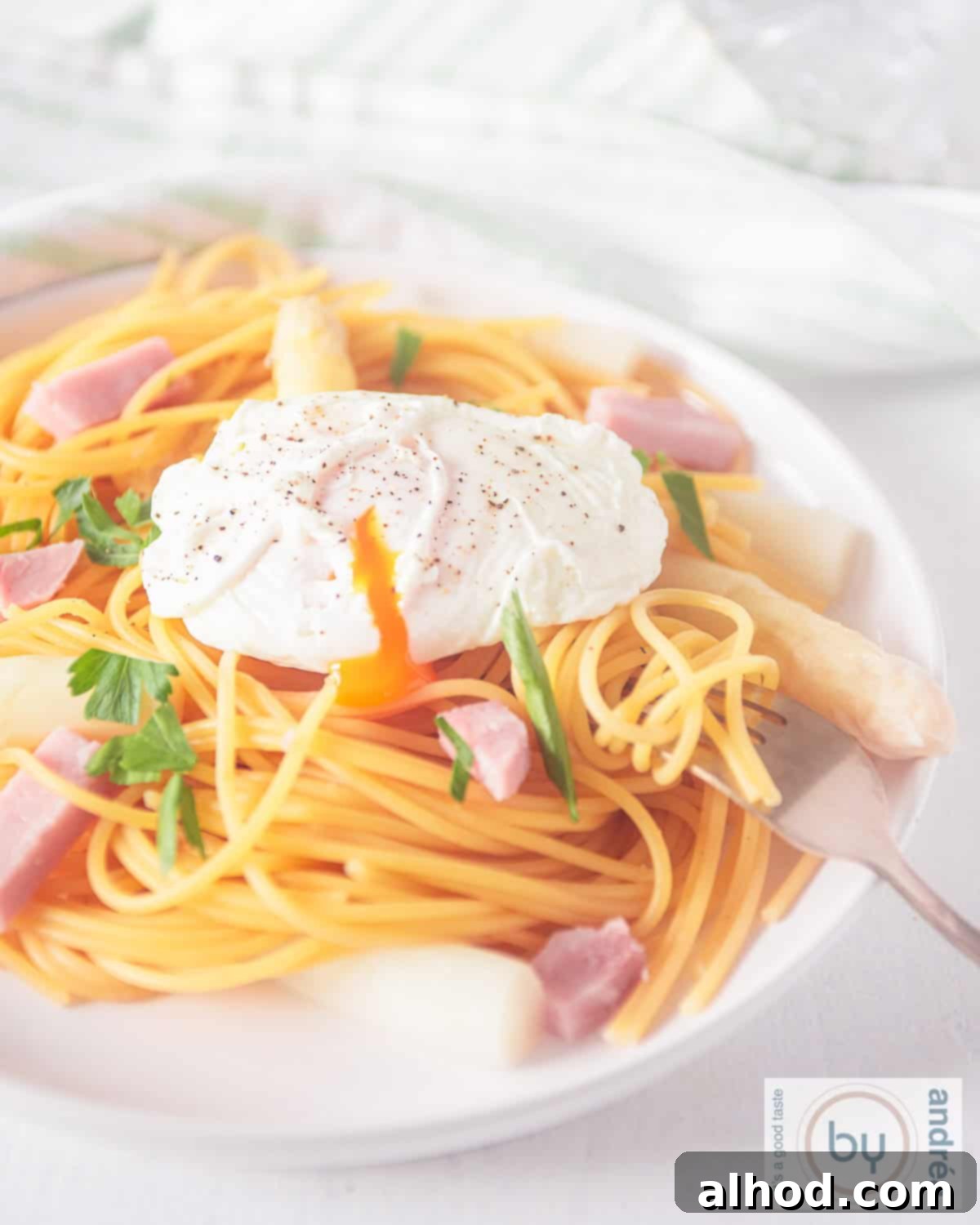 Effortless Asparagus Pasta with Poached Egg 2 A height picture of a white plate with spaghetti, asparagus, ham, and poached egg, garnished with fresh herbs.