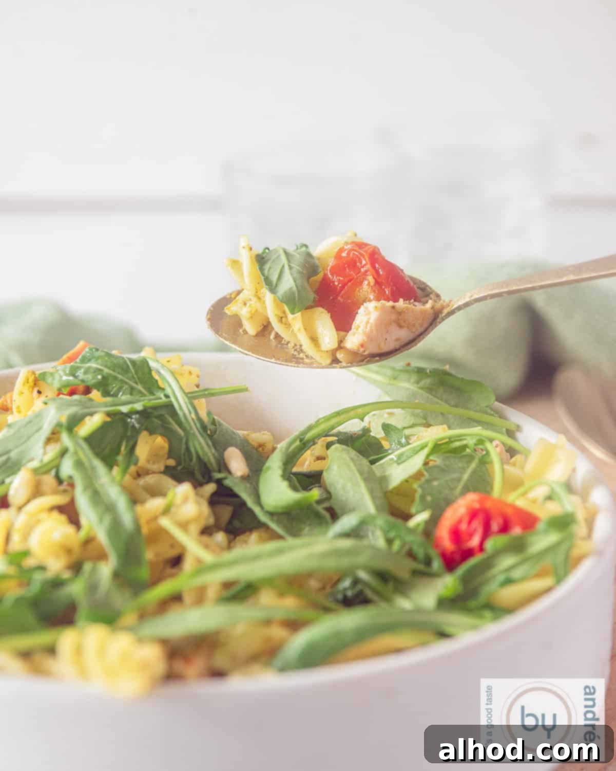 Smoky Pesto Chicken Pasta Salad 2 A close-up shot of a white bowl filled with Smoked Chicken Pesto Pasta Salad, showing fusilli pasta, pieces of smoked chicken, green arugula leaves, and red sun-dried tomatoes. A spoon is taking a bite.