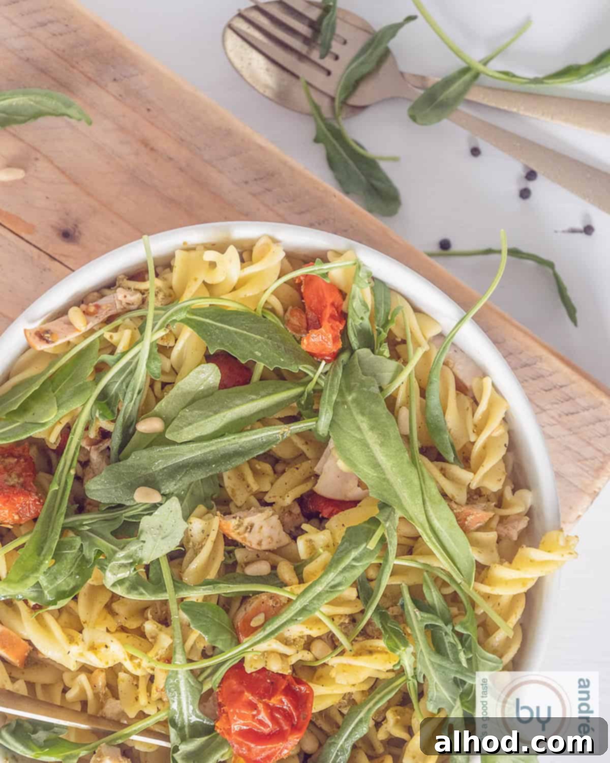 A photo from above of a white bowl overflowing with a colorful pasta salad featuring smoked chicken, green arugula, and rich pesto.