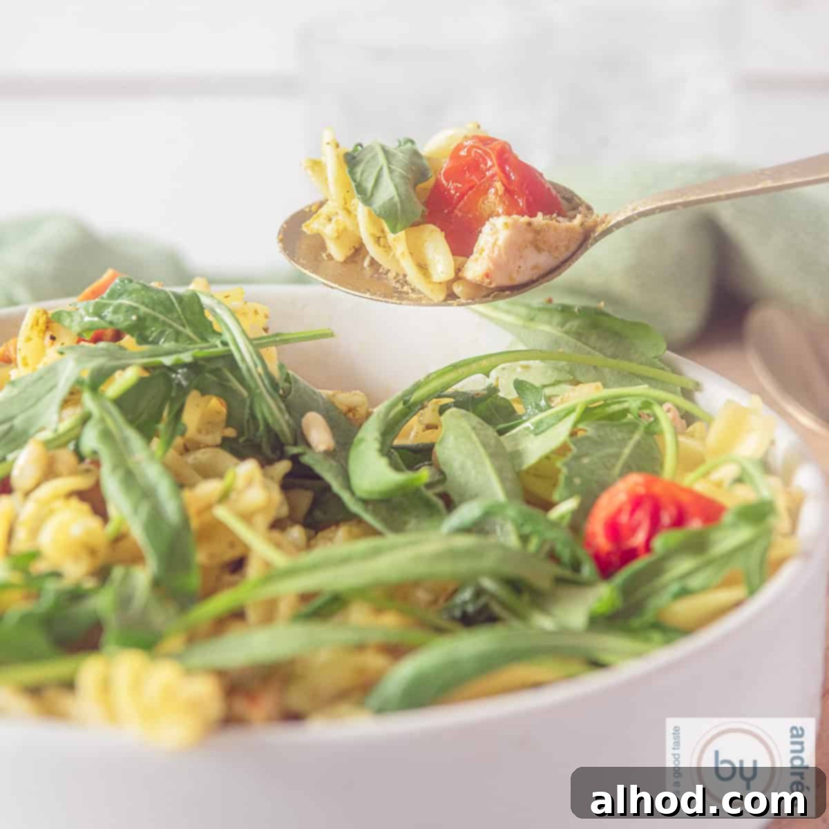Smoky Pesto Chicken Pasta Salad 4 A square photo showing part of a white bowl filled with fusilli pasta, arugula, sun-dried tomatoes, and green pesto sauce, making a delicious pasta salad.